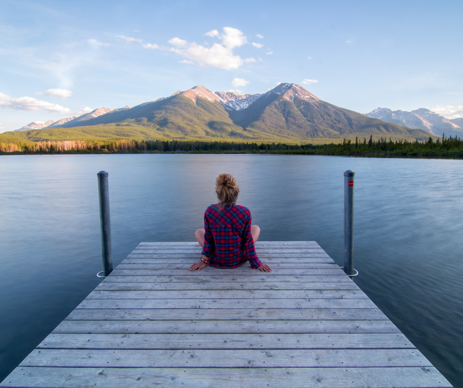 A woman sitting at the end of a dock taking in the view of the lake in front of her and mountains in the distance