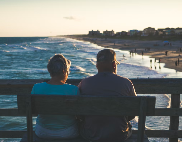 A photo of an older couple sitting on a pier. They are overlooking a beach at sunset.